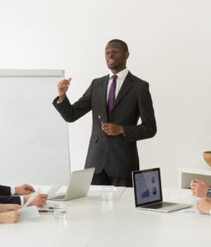 Confident african speaker business coach in suit giving presentation training sales team in office, black businessman speaking at seminar teaching people group presenting new marketing project goals