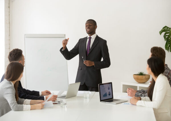 Confident african speaker business coach in suit giving presentation training sales team in office, black businessman speaking at seminar teaching people group presenting new marketing project goals